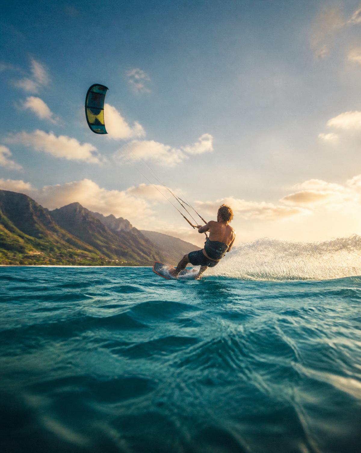Kitesurfista deslizándose sobre el mar turquesa con montañas costeras al fondo y luz de tarde